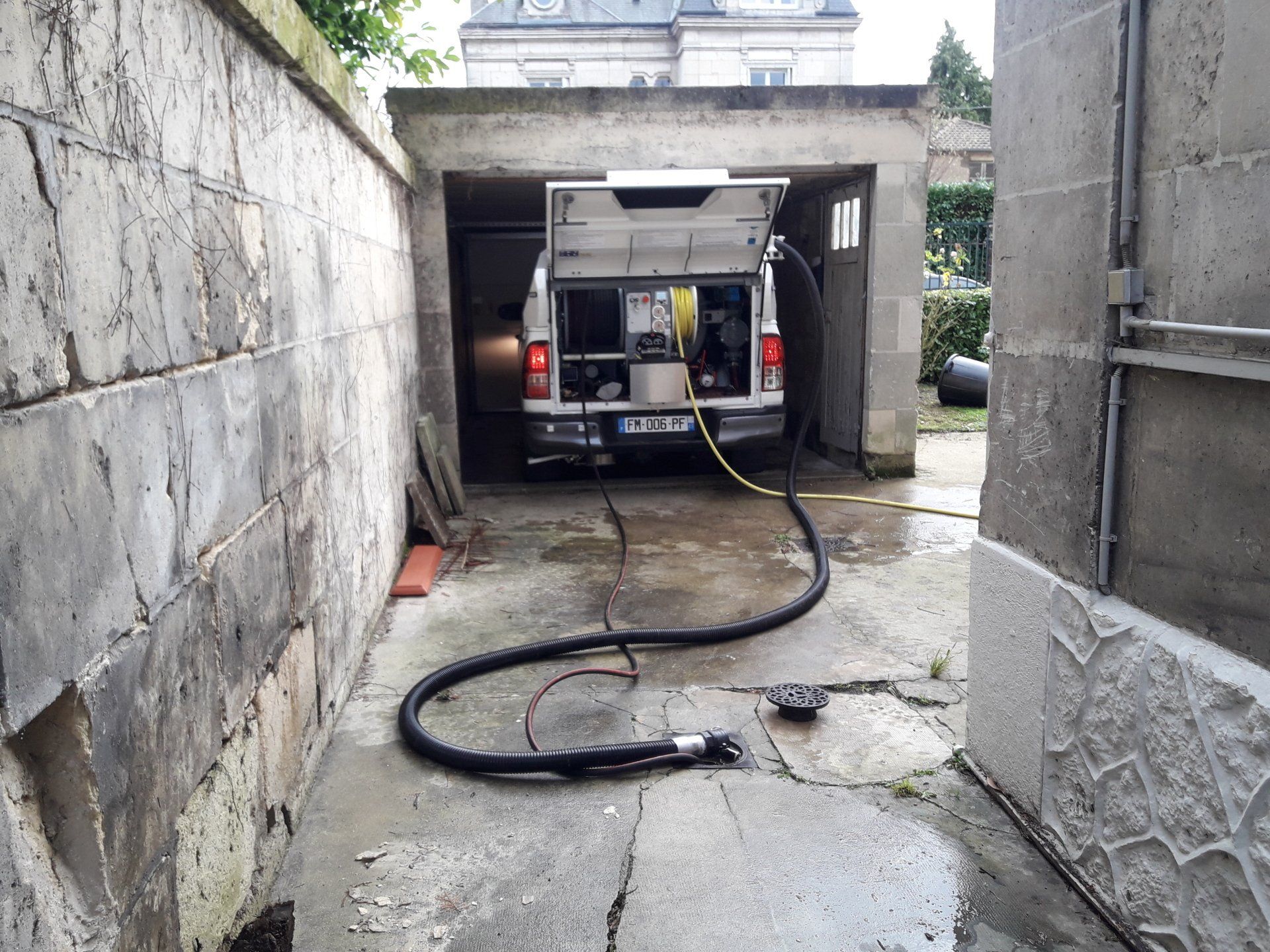 Camionnette garée dans une ruelle étroite en béton, tuyau d'arrosage tendu sur le sol humide en direction de l'entrée du garage.