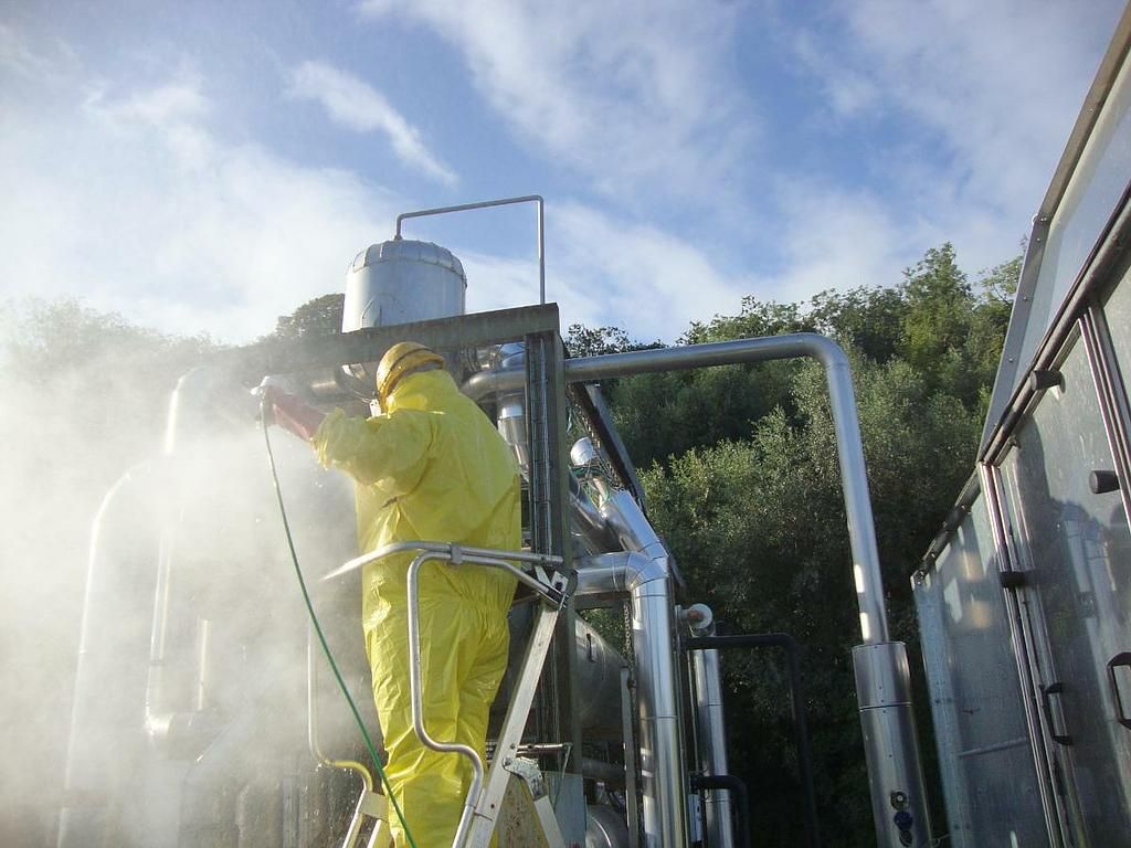 Ouvrier en combinaison de protection jaune nettoyant une structure industrielle à l'extérieur.