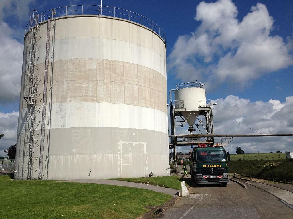 De grands réservoirs de stockage blancs bordent une route sous un ciel bleu, un véhicule est garé à proximité.