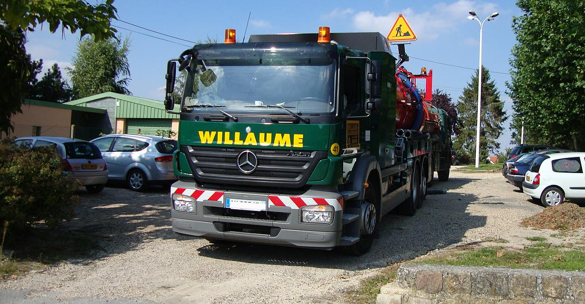 Camion vert sur un chemin de gravier dans un quartier résidentiel, avec des voitures garées à proximité.
