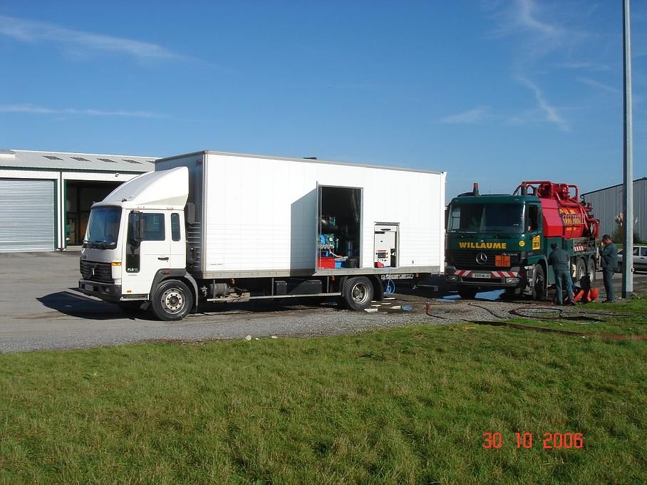Un camion blanc est stationné sur un terrain, sa porte latérale ouverte, à côté d'une dépanneuse verte et d'engins de chantier rouges.
