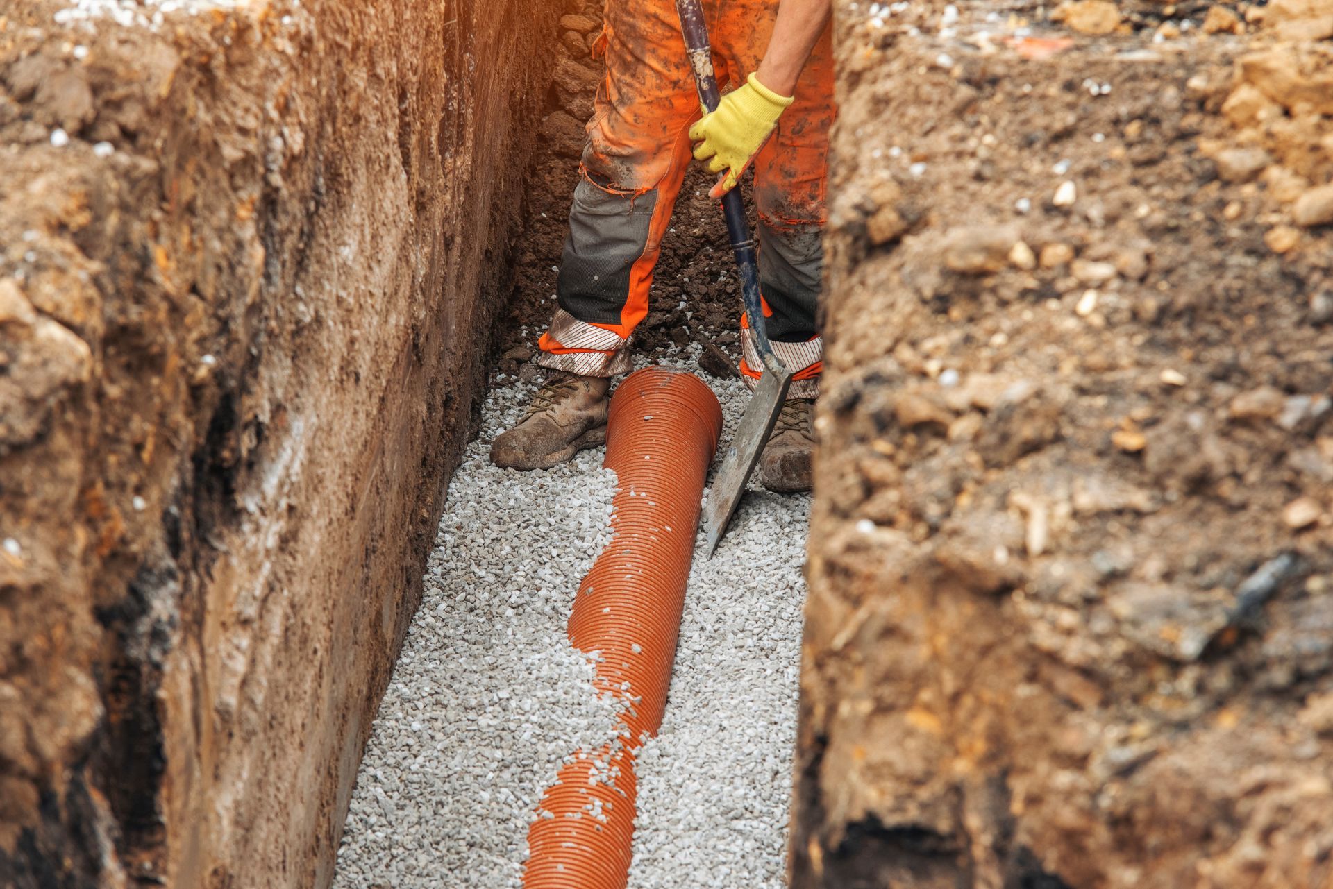 Ouvrier installant une canalisation souterraine orange dans une tranchée aux parois de gravier et de terre.
