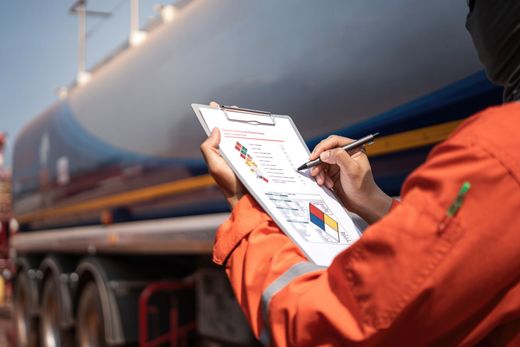 Un ouvrier en veste orange inspecte un camion avec un bloc-notes et un stylo.