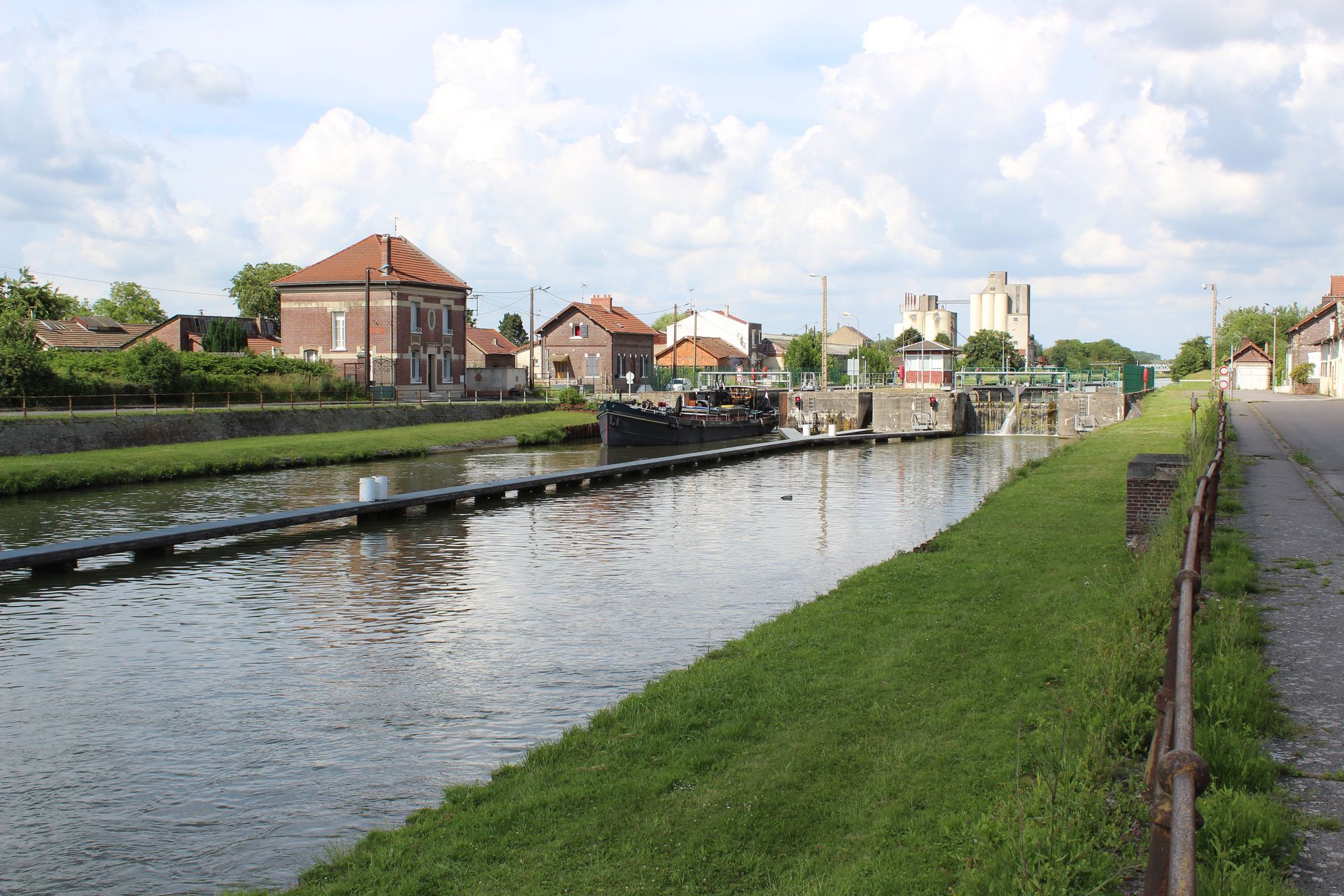 Canal aux eaux calmes, berge herbeuse, maisons et écluse sous un ciel partiellement nuageux.