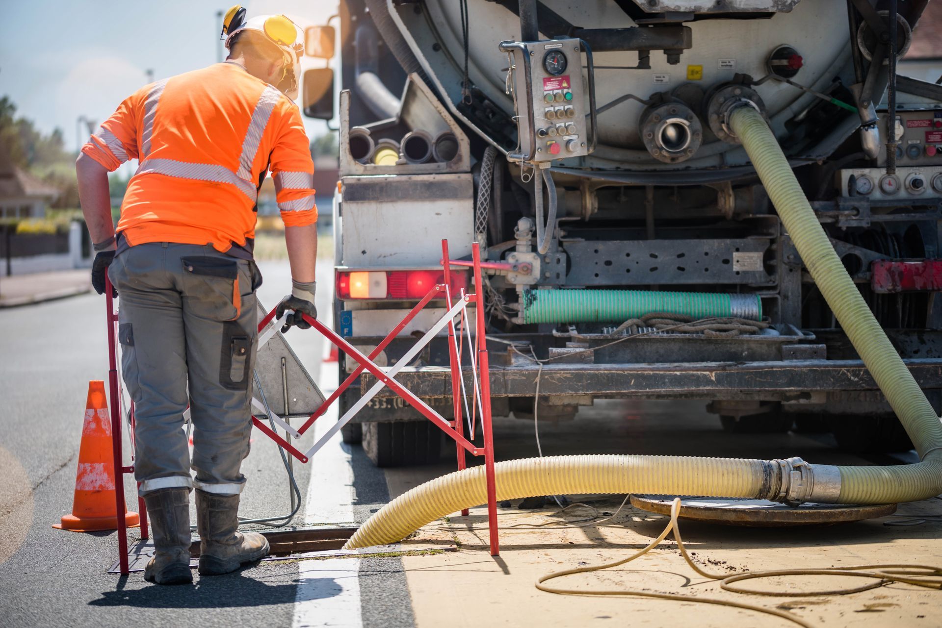 Un ouvrier en tenue de sécurité orange se tient à côté d'un camion de réparation routière et d'un tuyau d'arrosage dans une rue.