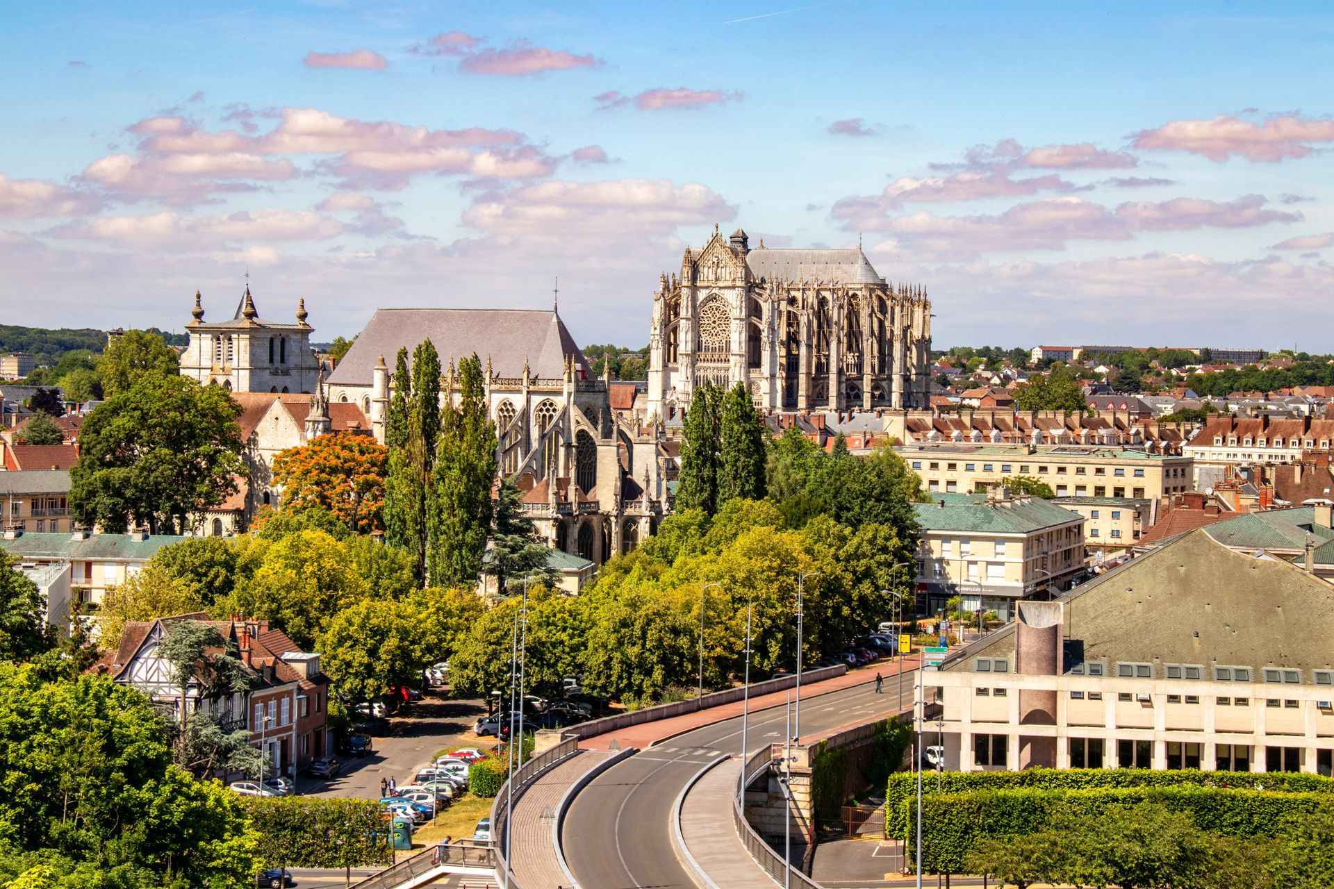 Paysage urbain avec une cathédrale, une route bordée d'arbres et des bâtiments historiques sous un ciel partiellement nuageux.