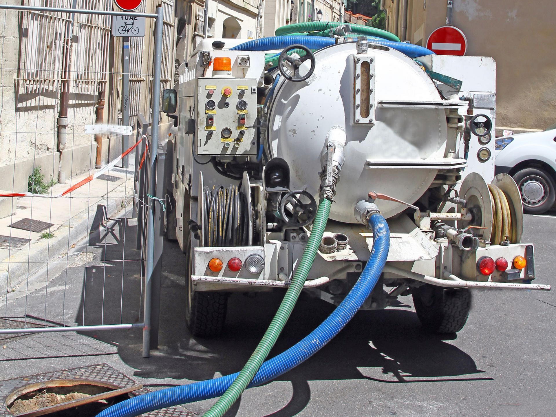 Camion de nettoyage des égouts avec tuyaux stationné dans une rue à côté d'un regard d'égout et de barrières de trottoir.