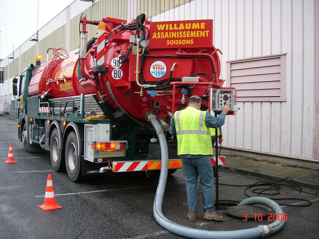 Un ouvrier nettoie un camion-citerne aspirateur rouge à l'extérieur d'un bâtiment à l'aide de tuyaux et de cônes orange.