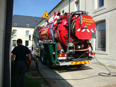 Un camion rouge de nettoyage des égouts, équipé de tuyaux, est stationné dans une rue étroite ; un employé se tient à côté.
