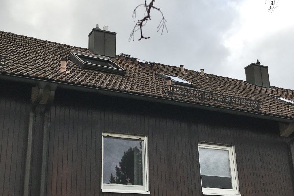 Braunes Gebäude mit Ziegeldach, Fenstern und zwei Schornsteinen unter bewölktem Himmel.