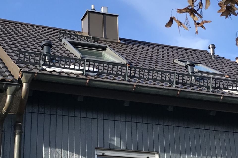Dunkles Dach mit Oberlichtern, Schornstein und Lüftungsöffnungen vor blauem Himmel.