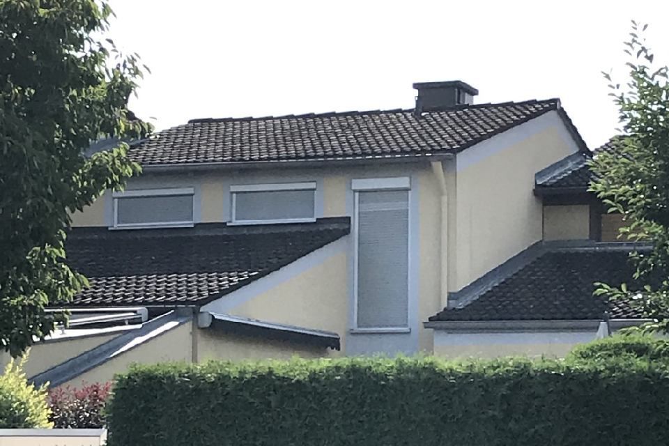 Beiges Haus mit dunkelgrauem Ziegeldach, drei Fenstern, Schornstein und einer grünen Hecke davor.