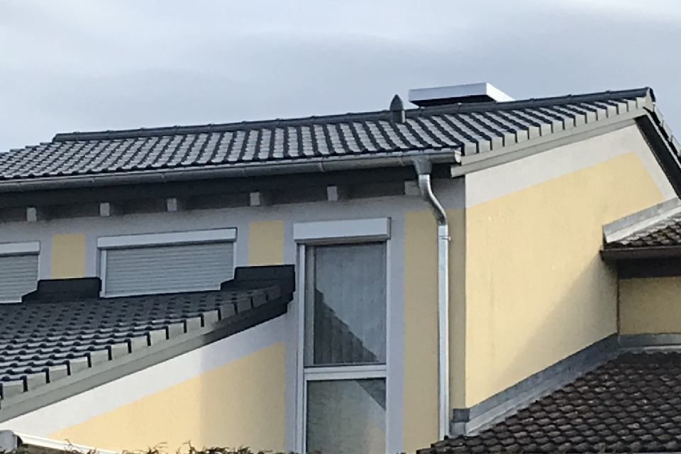 Gelbes Haus mit dunkelgrauem Ziegeldach und weißer Dachrinne unter bewölktem Himmel.