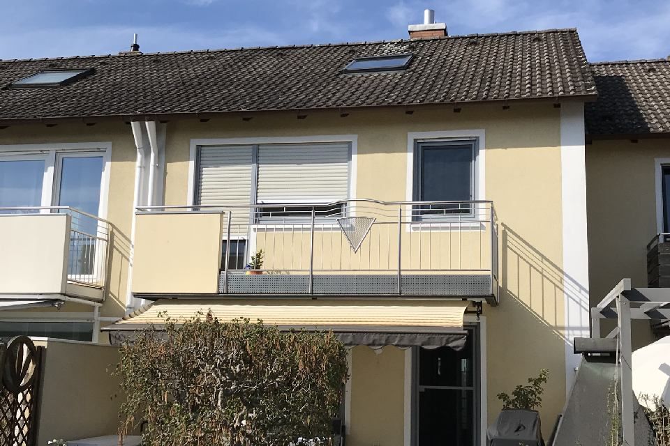Beiges Haus mit Balkon, Markise und Ziegeldach vor blauem Himmel.