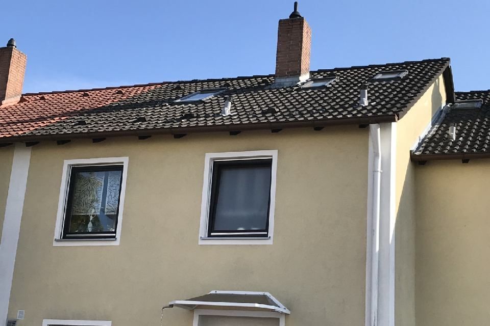 Ein beigefarbenes Gebäude mit dunklem Ziegeldach, zwei Fenstern und zwei Schornsteinen vor blauem Himmel.