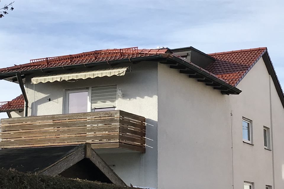 Zweistöckiges Gebäude mit rotem Ziegeldach, Balkon und beiger Markise an einem sonnigen Tag.
