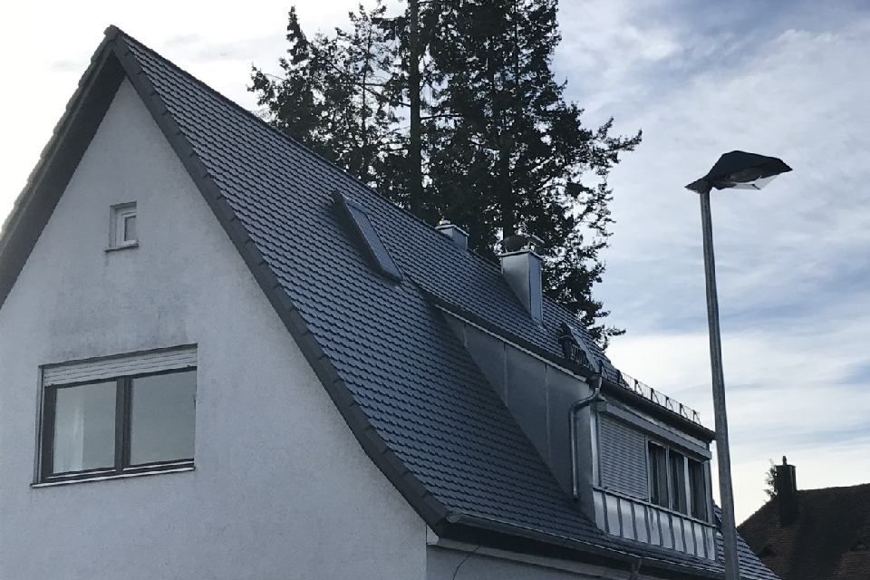 Weißes Haus mit dunkelgrauem Ziegeldach, Dachgaube und Straßenlaterne vor bewölktem Himmel.