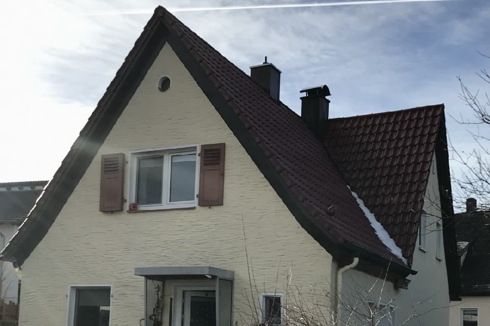 Beiges Haus mit rotem Dach, dunklen Zierelementen, weißen Fenstern und Fensterläden. Schornstein und bedeckter Himmel sind sichtbar.