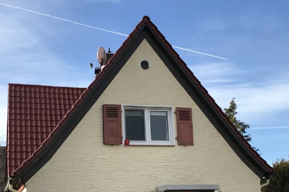 Giebel eines A-förmigen Hauses mit braunen Fensterläden, Fenster und rotem Ziegeldach vor blauem Himmel.