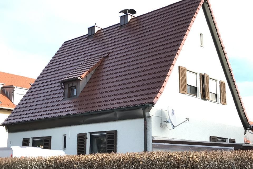 Weißes Haus mit braunem Ziegeldach, kleinen Fenstern, Fensterläden und einer Satellitenschüssel.