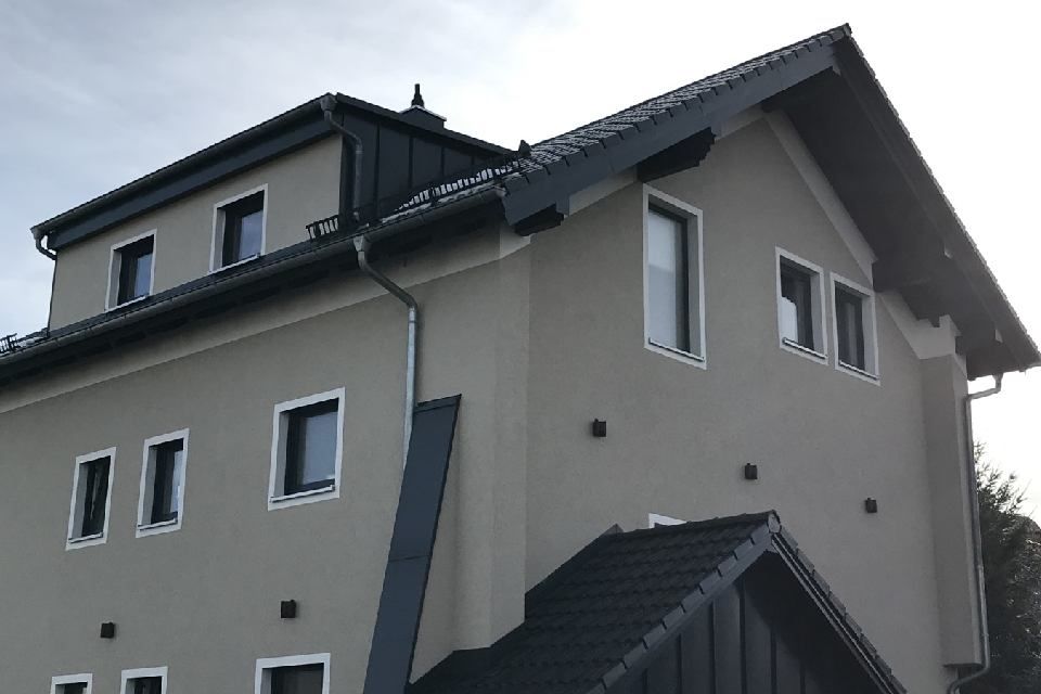 Zweistöckiges Haus mit grauen Stuckwänden, dunklem Dach und weiß umrandeten Fenstern.