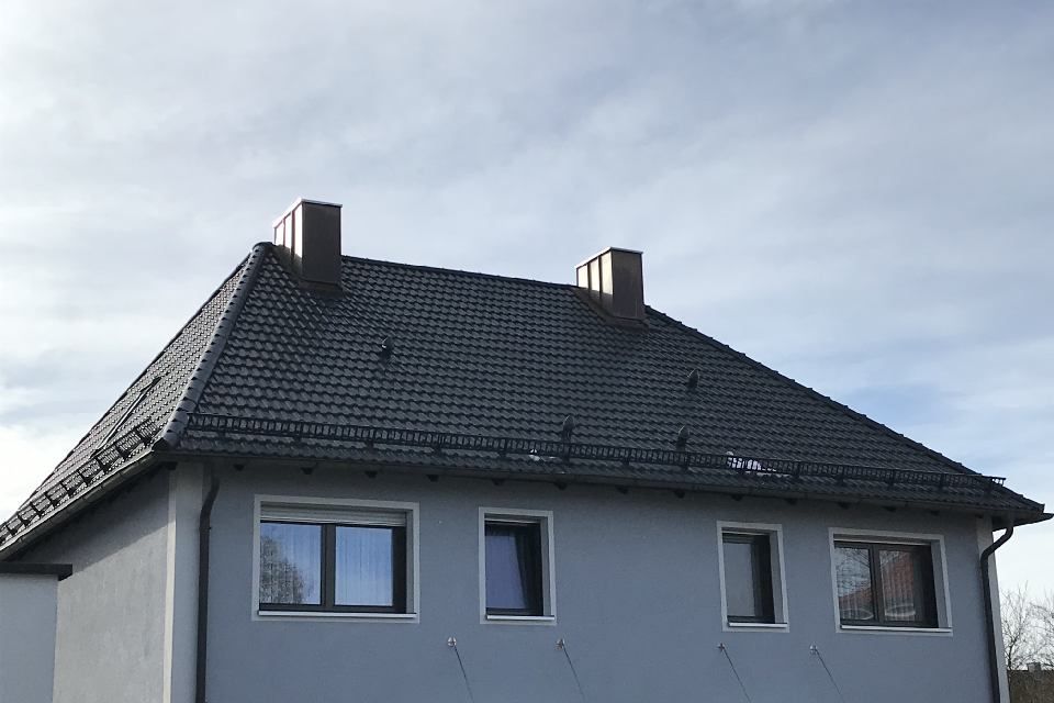Graues Haus mit dunkelgrauem Ziegeldach, zwei Schornsteinen und vier Fenstern vor einem bewölkten Himmel.