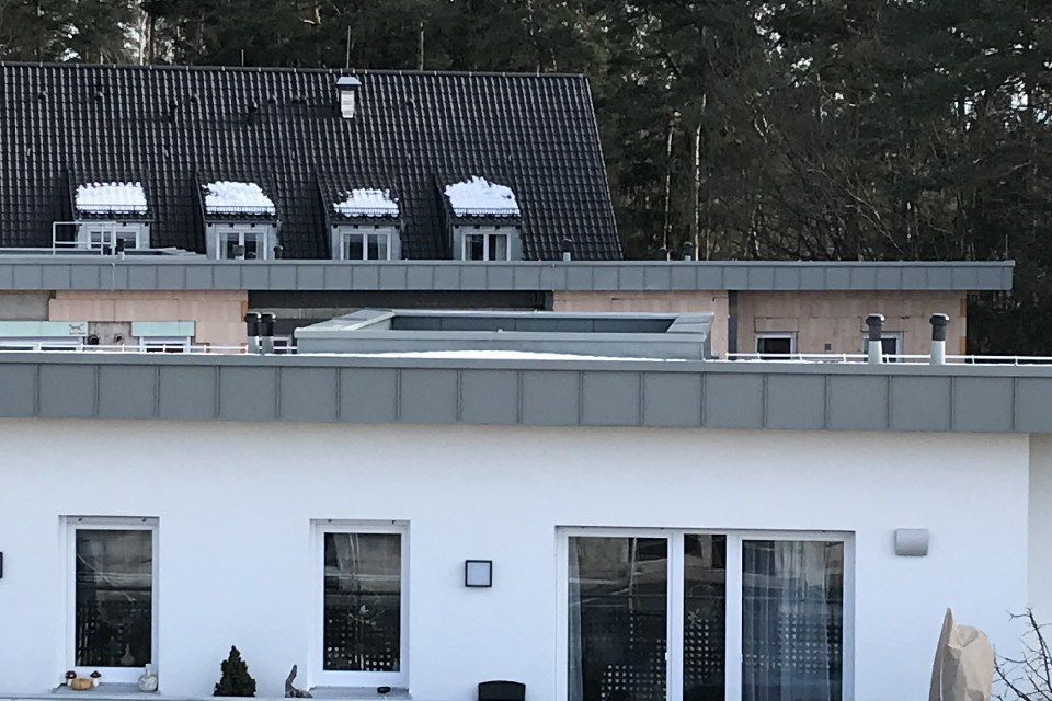 Weißes Gebäude mit mehreren Fenstern und Flachdach, Schnee auf dem Dach einer Dachgaube, Bäume im Hintergrund.