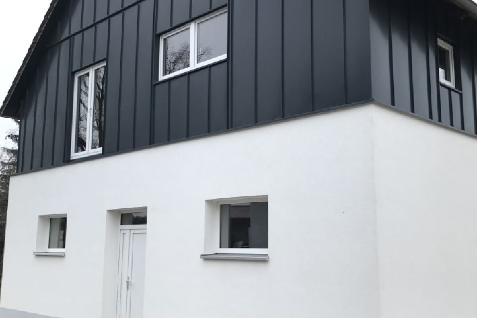 Zweistöckiges Haus mit schwarzer Holzverkleidung im oberen Bereich und weißem Stuck im unteren Bereich. Mehrere Fenster.