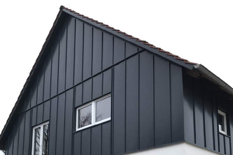 Dunkelgraue, vertikale Holzverkleidung an einem Haus mit weiß gerahmten Fenstern vor weißem Himmel.