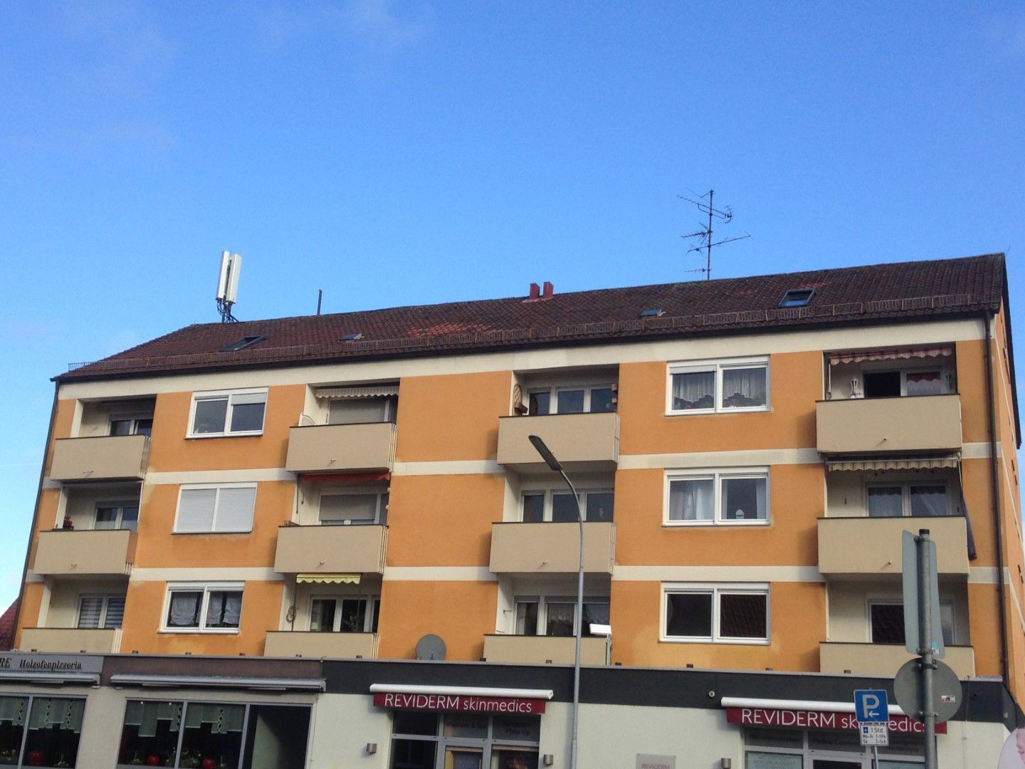 Orangefarbenes Wohnhaus mit Balkonen, braunem Dach, darunter Ladenfronten, klarer blauer Himmel.