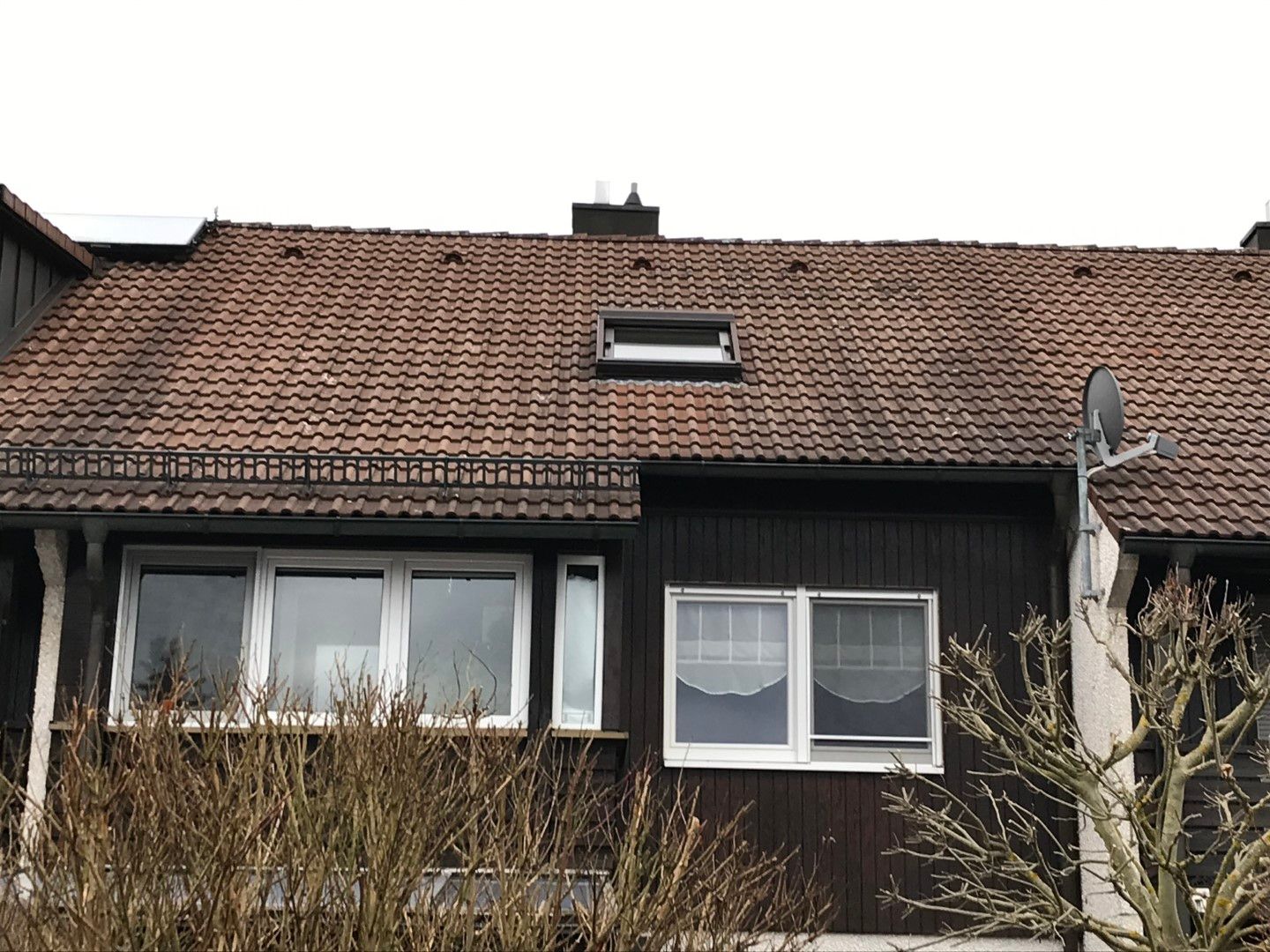 Ein Gebäude mit braunem Dach und mehreren Fenstern, darunter ein Oberlicht. Eine Satellitenschüssel und kahle Äste sind zu sehen.