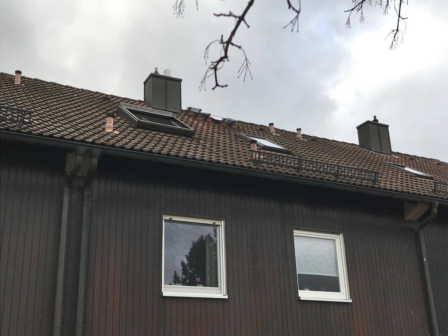 Braunes Gebäude mit Ziegeldach, Fenstern und zwei Schornsteinen unter bewölktem Himmel.