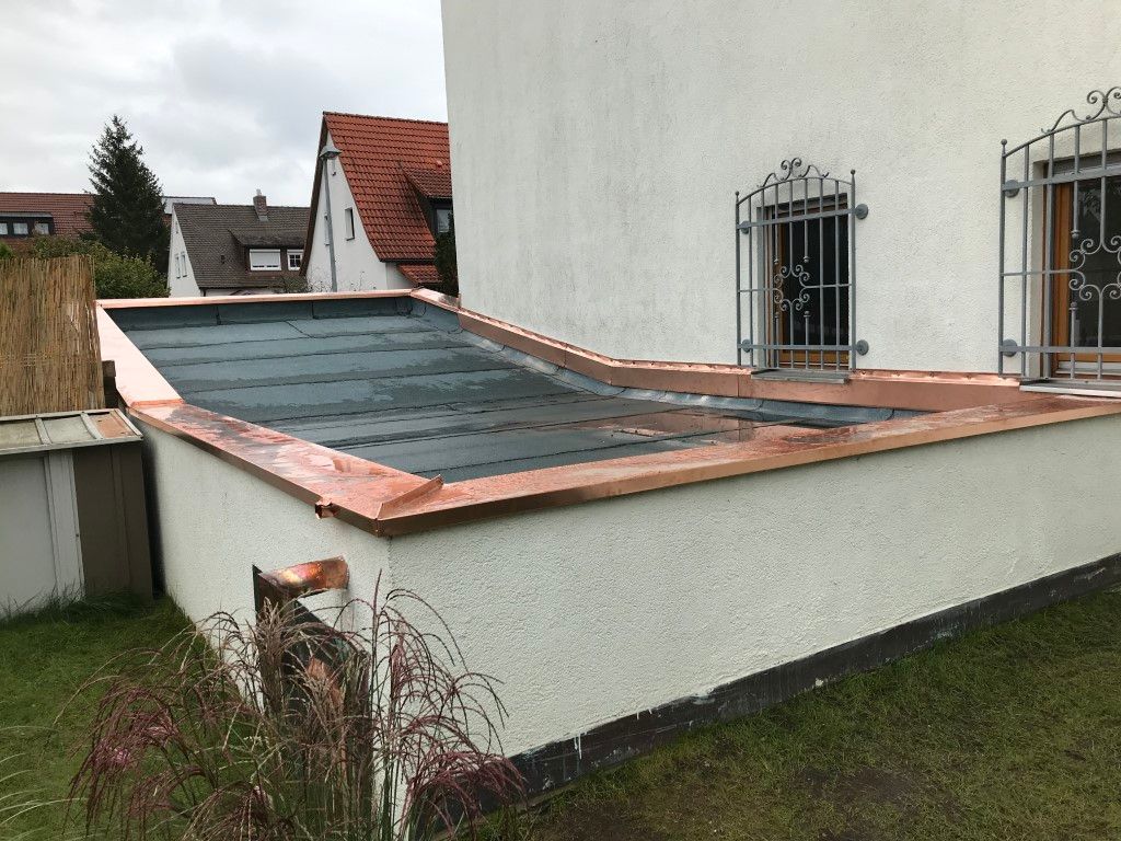 Flachdach mit Kupferverkleidung auf einem weißen Gebäude, Gras im Vordergrund, Häuser im Hintergrund.