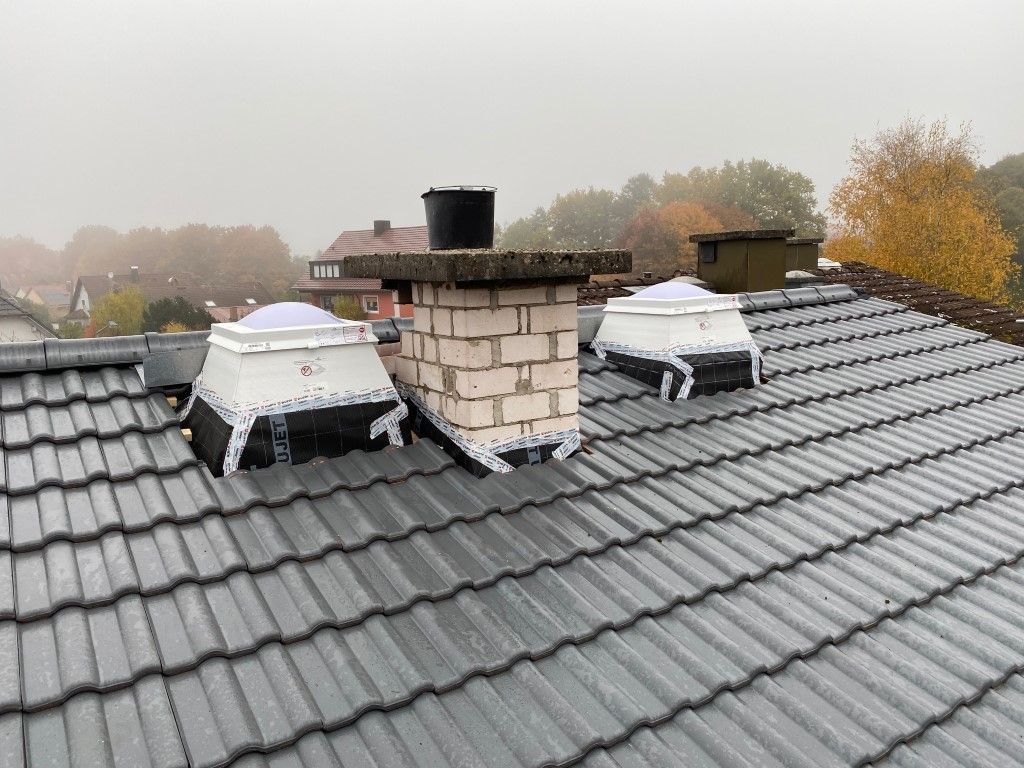 Ein graues Ziegeldach mit einem gemauerten Schornstein und zwei mit schwarzem Stoff verkleideten Dachfenstern. Bewölkter Tag.