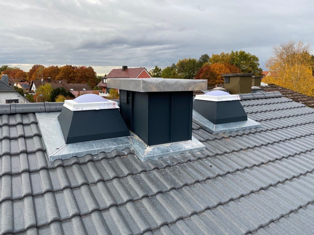 Drei dunkelgraue Dachlüfter auf einem grauen Ziegeldach, mit Blick auf herbstliche Bäume und bewölkten Himmel.