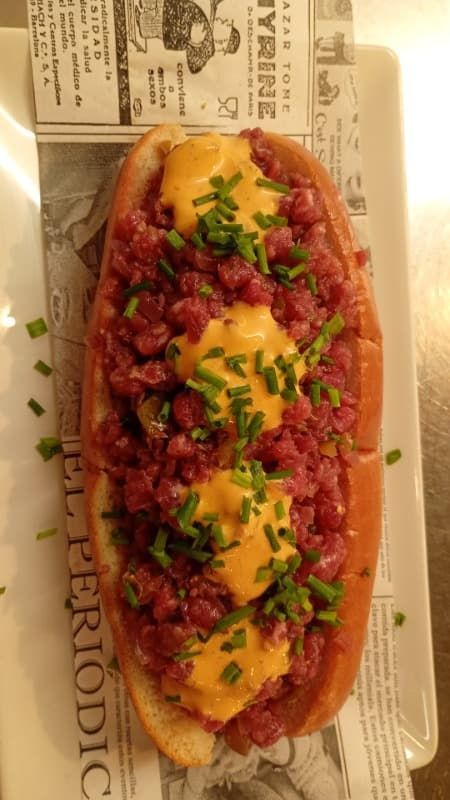 Perrito caliente cubierto con trocitos de tocino, queso derretido y cebollino en una bandeja con una tarjeta de menú al lado.