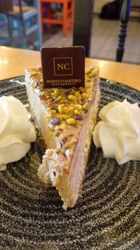 Una porción de pastel de pistacho sobre un plato negro con cucharadas de crema batida, coronada con un cuadrado de chocolate con el logo.