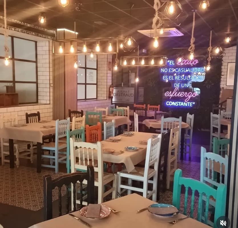 Interior acogedor del restaurante con sillas en tonos pastel, lámparas colgantes, mesas puestas y un letrero de neón en la pared.