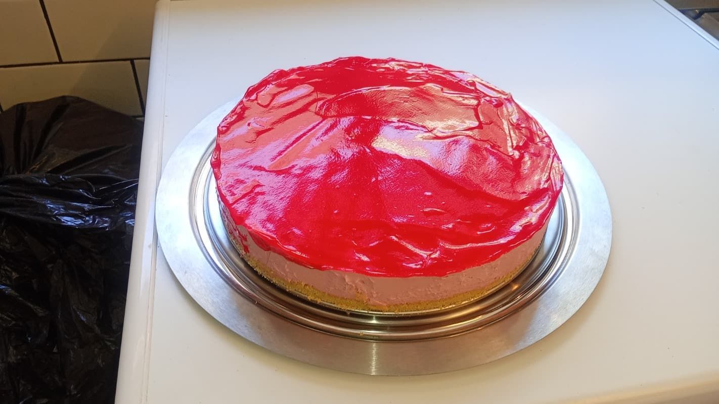 Pastel redondo con glaseado rojo sobre una bandeja plateada en una mesa blanca.