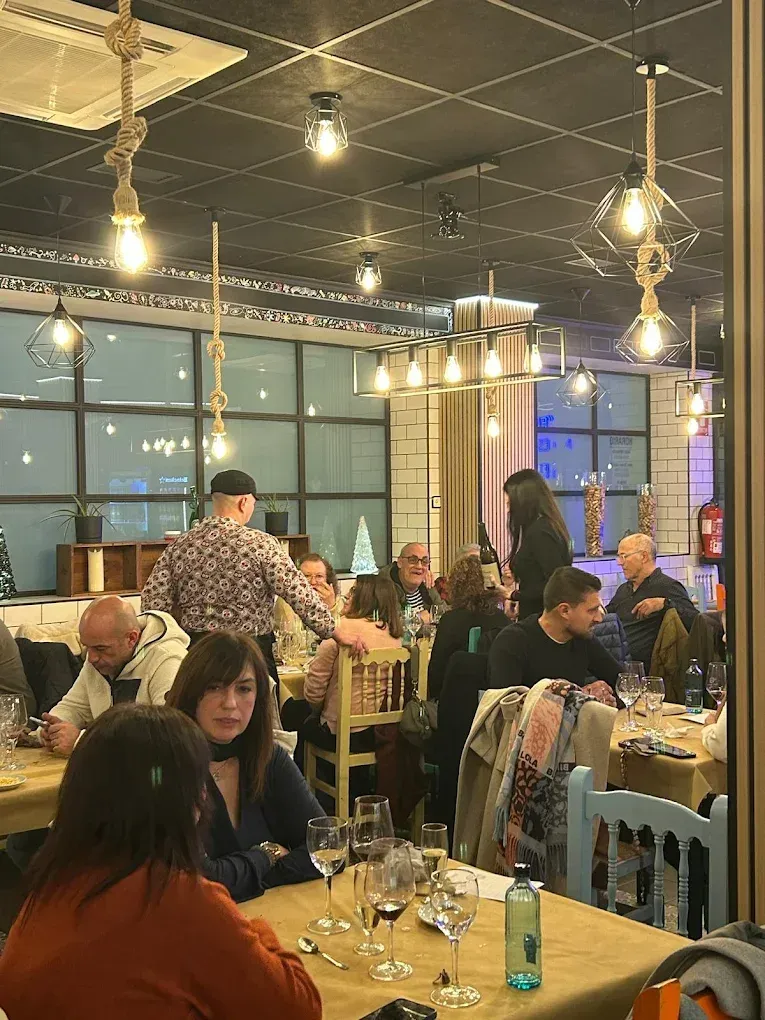 Interior del restaurante: gente sentada en mesas, algunos conversando. Lámparas colgantes, fondo de ventana.