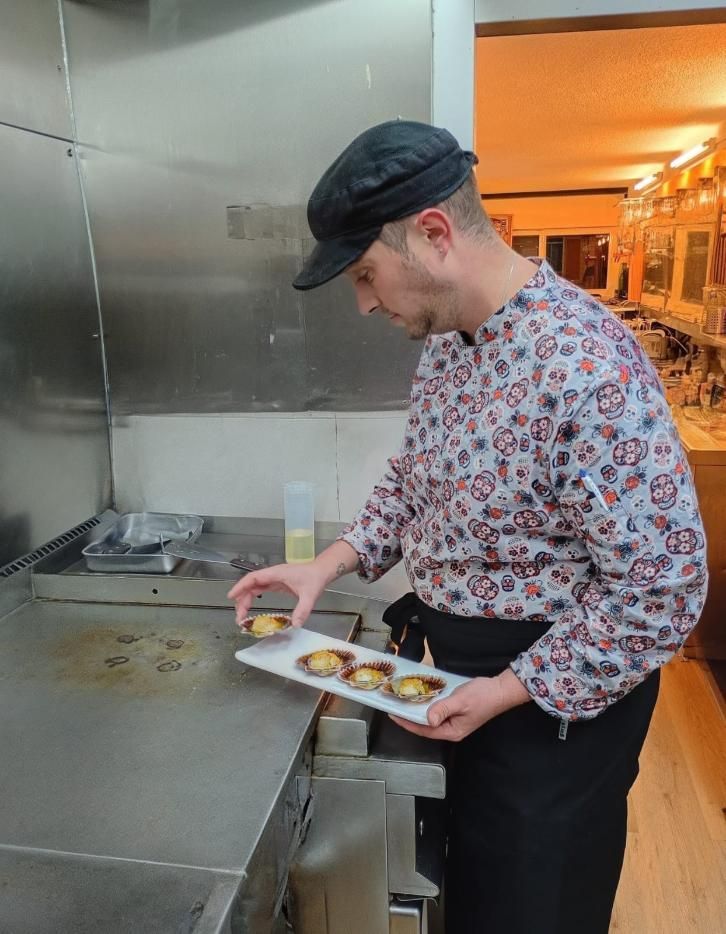 Chef sirve tartaletas en un plato blanco cerca de una estufa de metal. Lleva sombrero, camisa estampada y delantal negro.