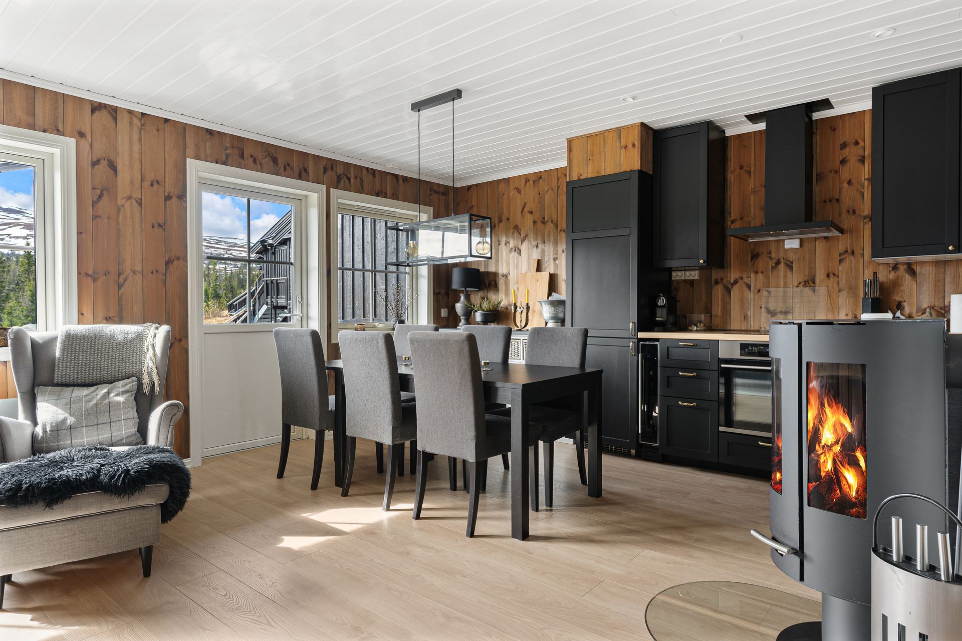 Cozy cabin interior with wooden walls, a dark kitchen, dining table, and fireplace.
