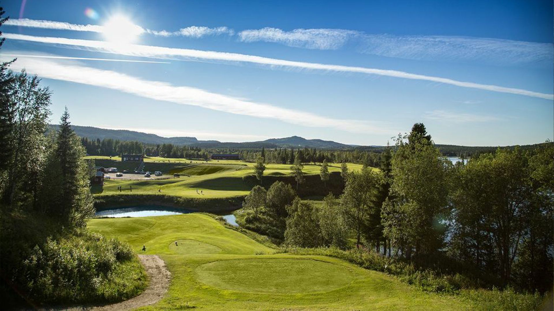 Green golf course with blue sky, trees, and lake under bright sun.