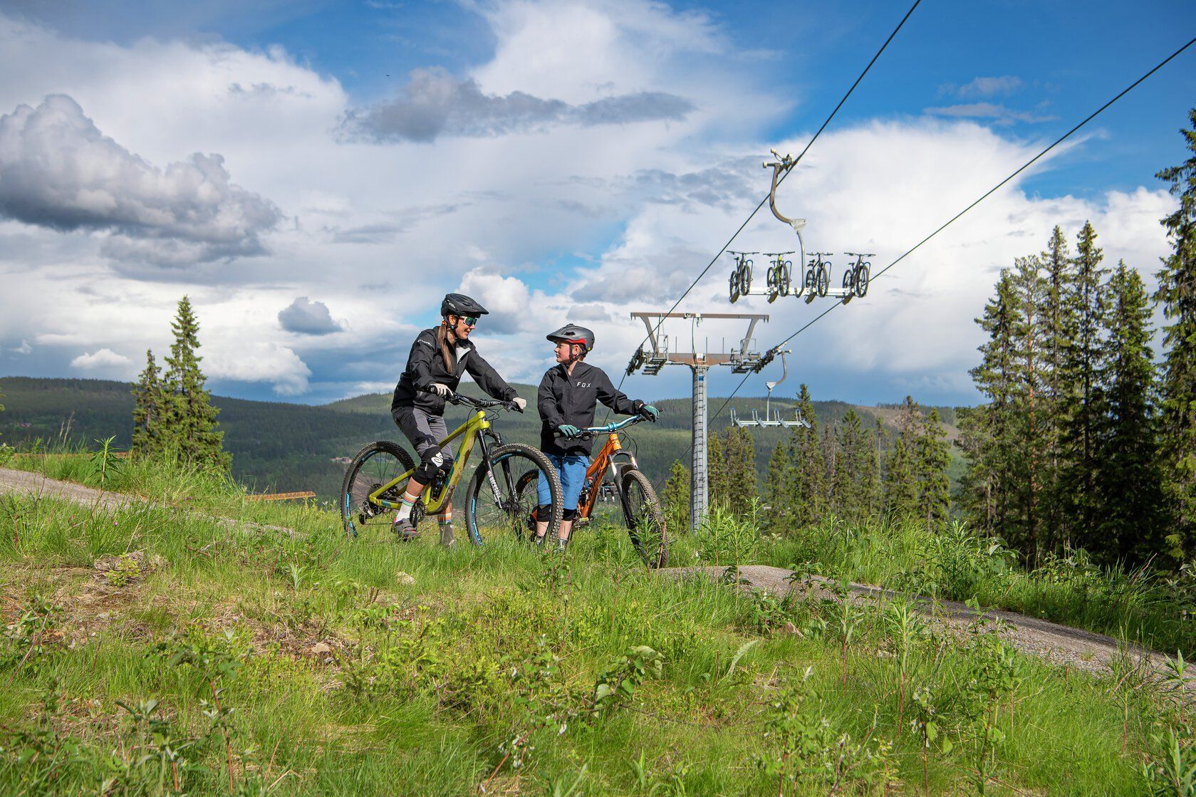 Two mountain bikers, ready to ride, near a chairlift. Green grass, cloudy sky, and forest background.