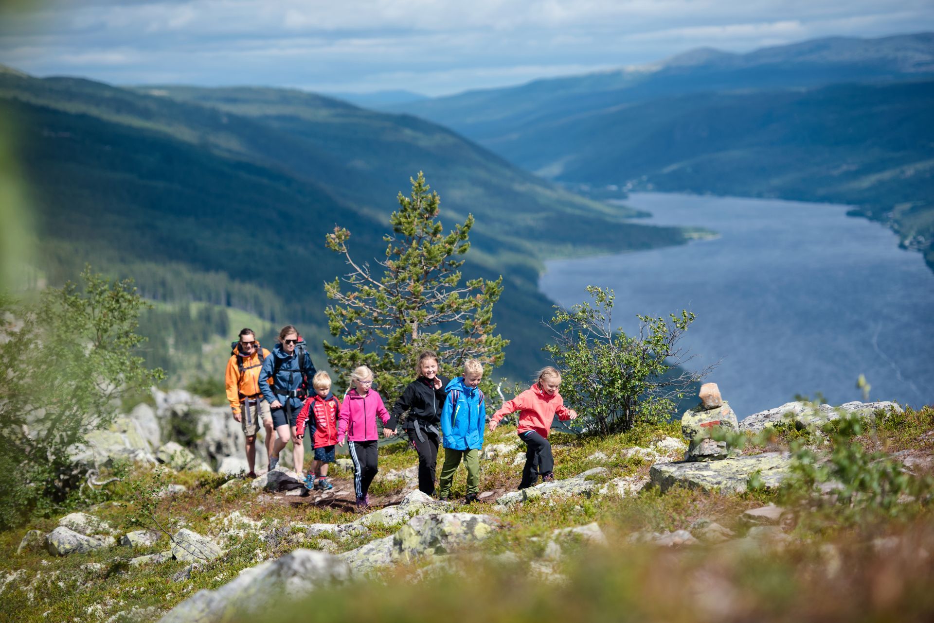 Family hiking on a rocky trail overlooking a lake and mountains under a blue sky.