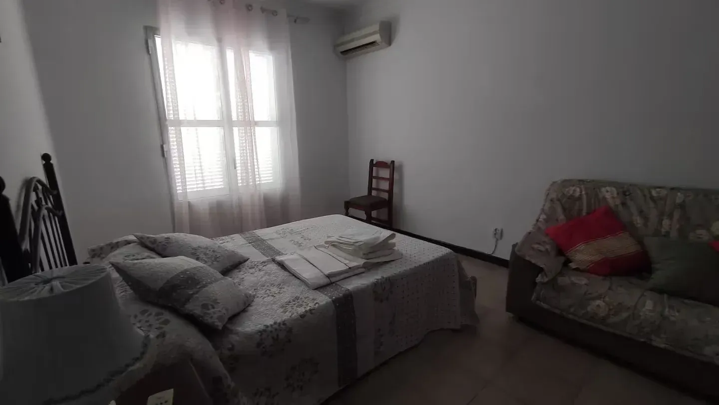 Dormitorio con cama, sofá, sillón, ventana y aire acondicionado. Gris, blanco y marrón.