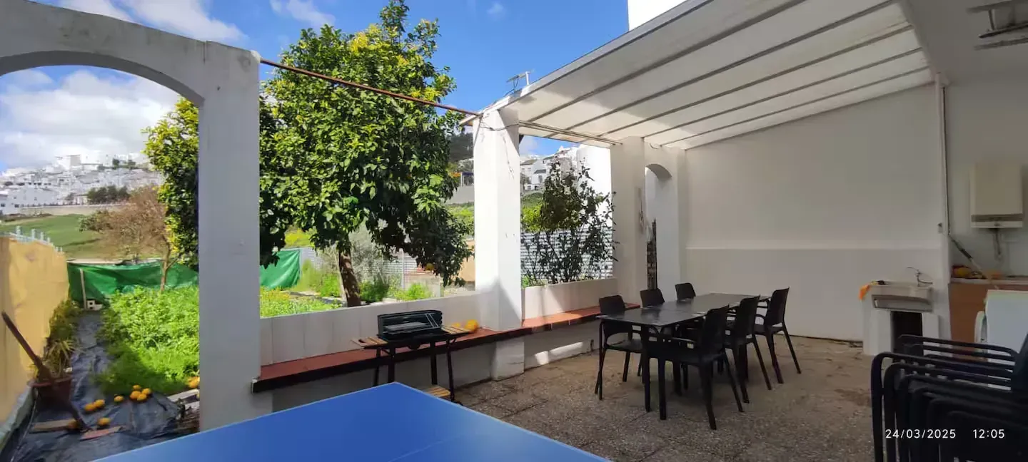 Patio exterior con mesa de comedor y barbacoa bajo un toldo. Con vistas a un jardín arbolado y verde.