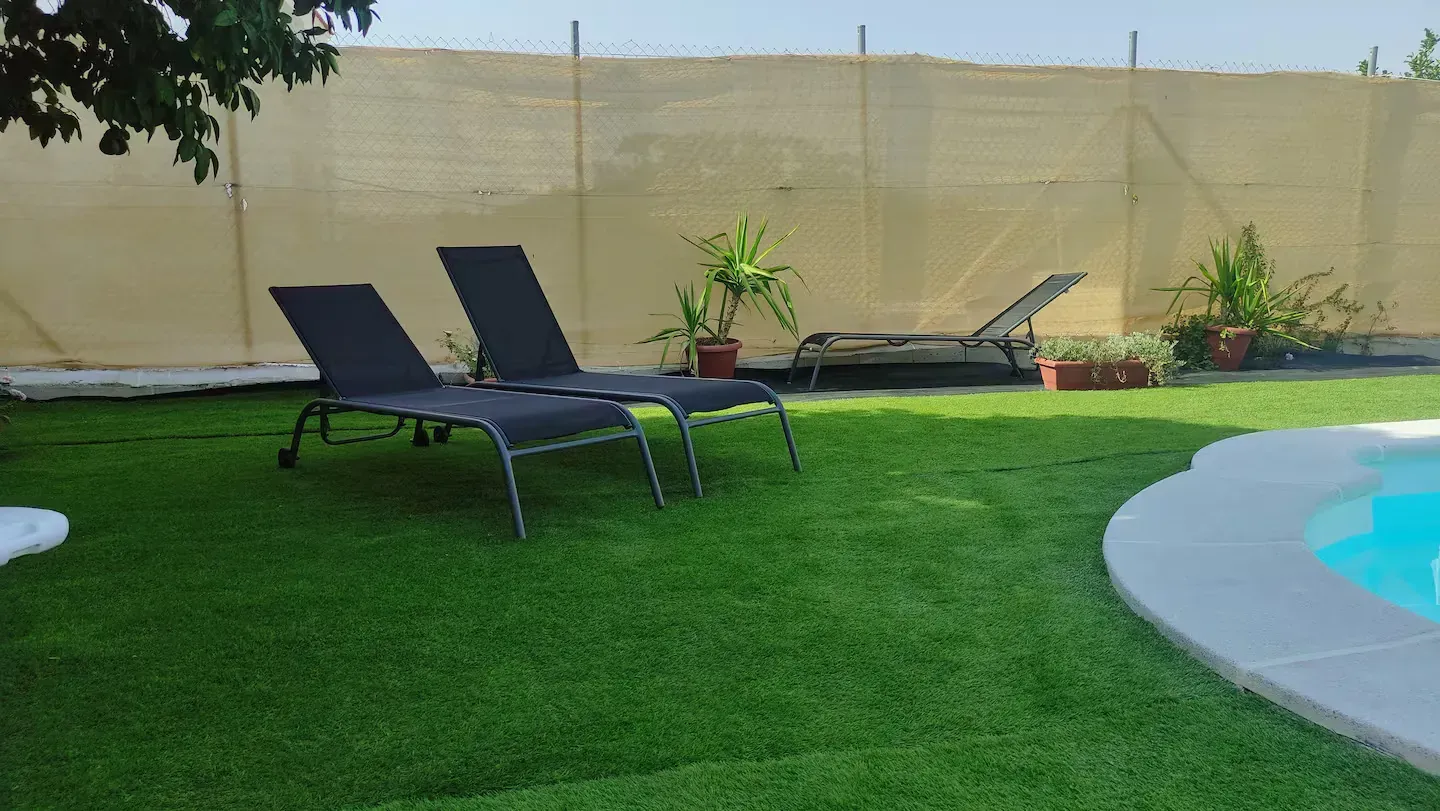 Tumbonas sobre césped artificial verde junto a una piscina. Valla beige al fondo con plantas en maceta.