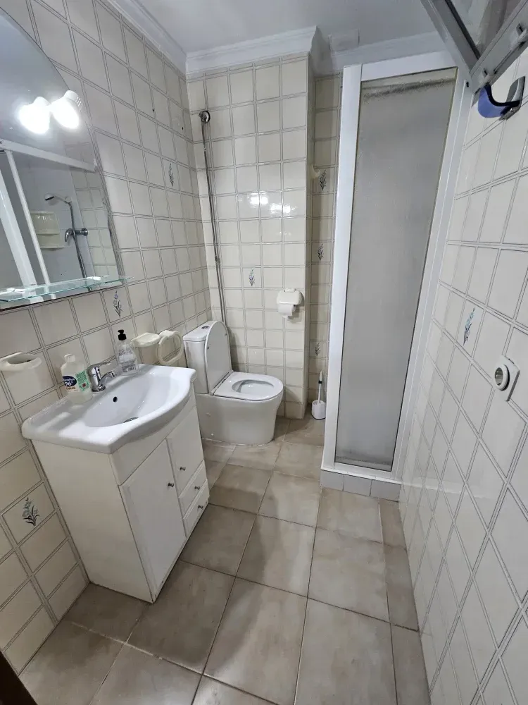 Baño con lavabo, inodoro y ducha blancos. Paredes y suelo de azulejos. Espejo sobre el lavabo.