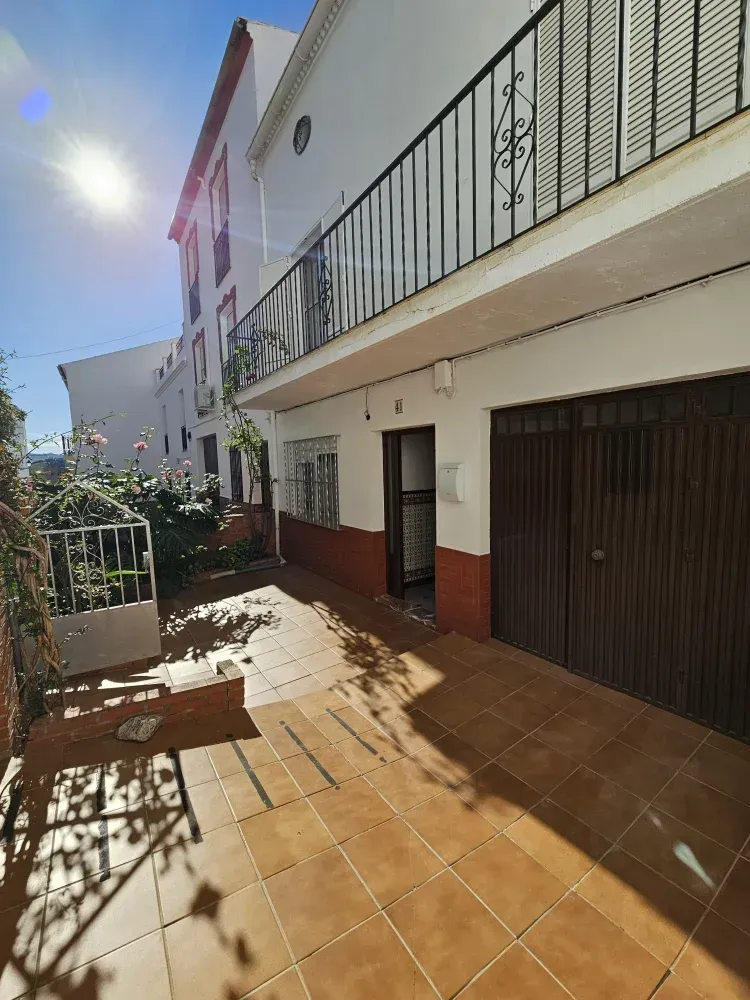 Exterior de casa adosada con patio de baldosas marrones, puerta de garaje, balcón y sol brillante.